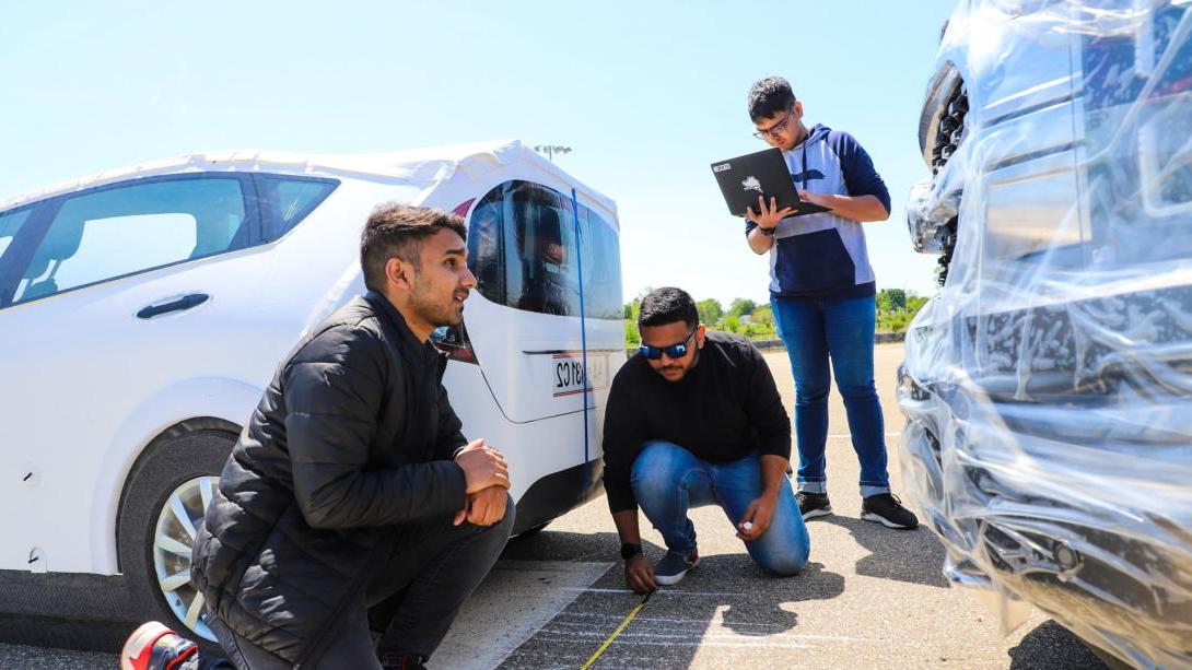 Three Kettering students measure markings on a road at the Mobility Research Center using measuring tape. They stand between two cars.